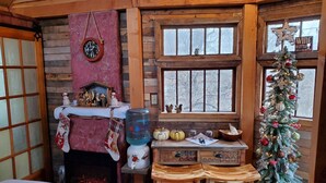 Ein gemütliches Blockhaus-Interieur mit Holzvertäfelung, Kamin, Weihnachtsbaum und Fenster mit Blick auf kahle Bäume.