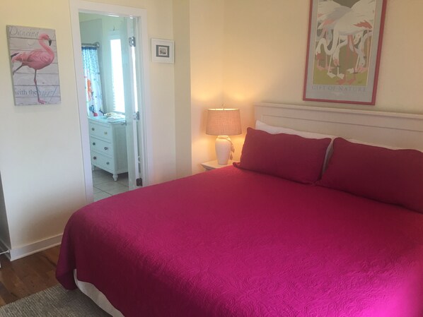 A bedroom with a large bed, a nightstand with a lamp, a dresser, and a door leading to another room.