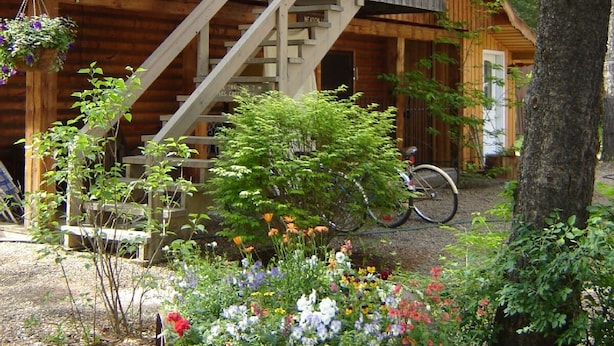 A wooden cabin with a staircase leading to a balcony, surrounded by a garden with various plants and flowers. - Martins Vacation Rental Log Cabin (Hungry Horse)