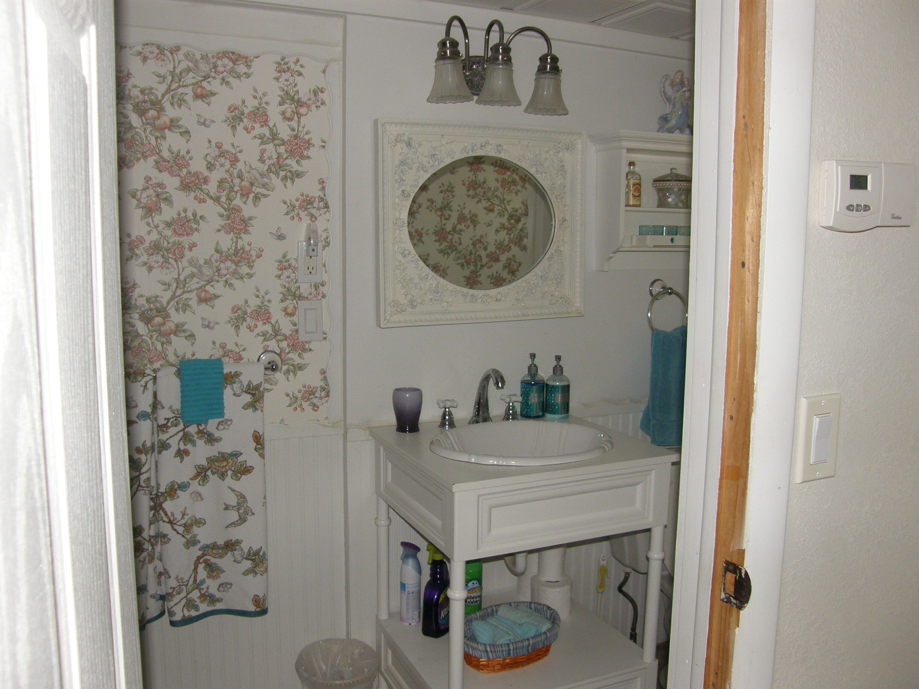 Uma casa de banho com uma pia branca, um espelho acima dela e um papel de parede floral. Existem duas luminárias acima do espelho e um porta-toalhas à direita.