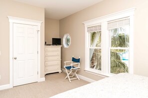 A bedroom with a bed, a dresser, a chair, a television, and a window with a view of palm trees.