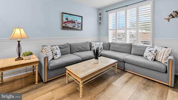 Ein Wohnzimmer mit grauem Ecksofa, einem Holztisch, einer Stehlampe und einem Deckenventilator. Ein Fenster mit Jalousien und ein gerahmtes Bild sind an der Wand zu sehen.