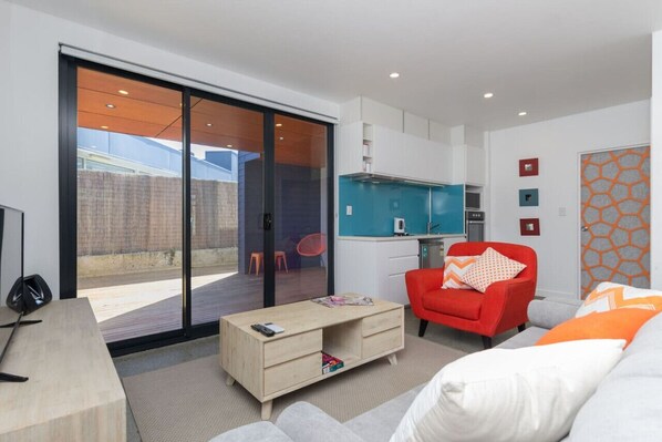 A modern living room with a sliding glass door leading to a patio, a small kitchenette, and a red armchair.