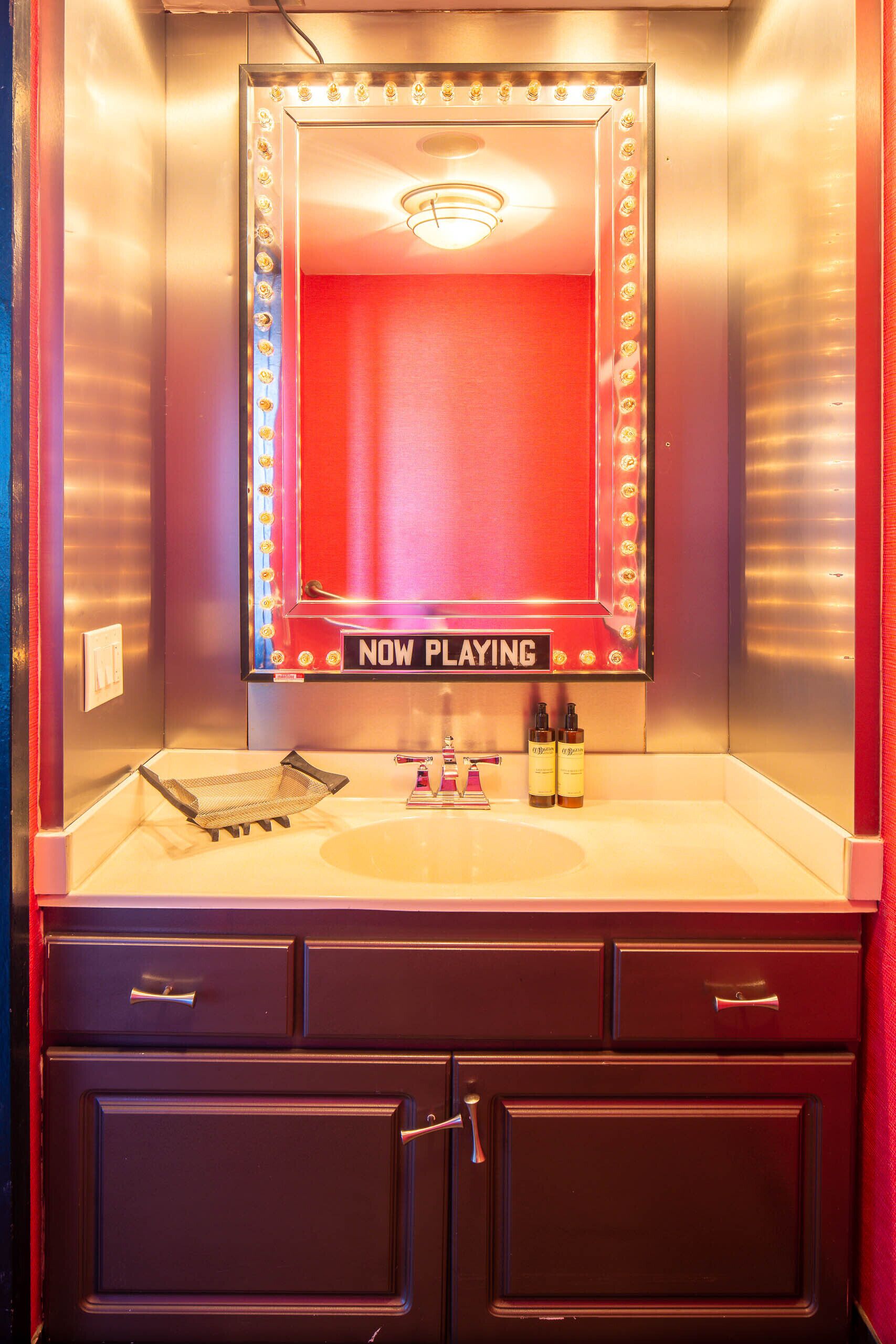 A bathroom vanity with a mirror, vanity lights, and a 'NOW PLAYING' sign.