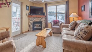 Ein gemütliches Wohnzimmer mit Kamin aus Stein, Deckenventilator, großem Fenster mit Blick auf eine verschneite Landschaft, bequemer Couch und Holztisch.