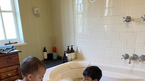 Zwei Kinder spielen in einer Badewanne mit einem Holzkabinett und einer gefliesten Wand im Hintergrund.