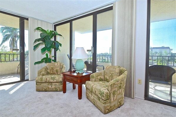 A room with two armchairs, a small table, and a large plant. The room has large windows and a view of palm trees.