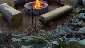Une cabane en bois rustique, avec un foyer au premier plan, entourée de troncs d’arbres et de rochers.