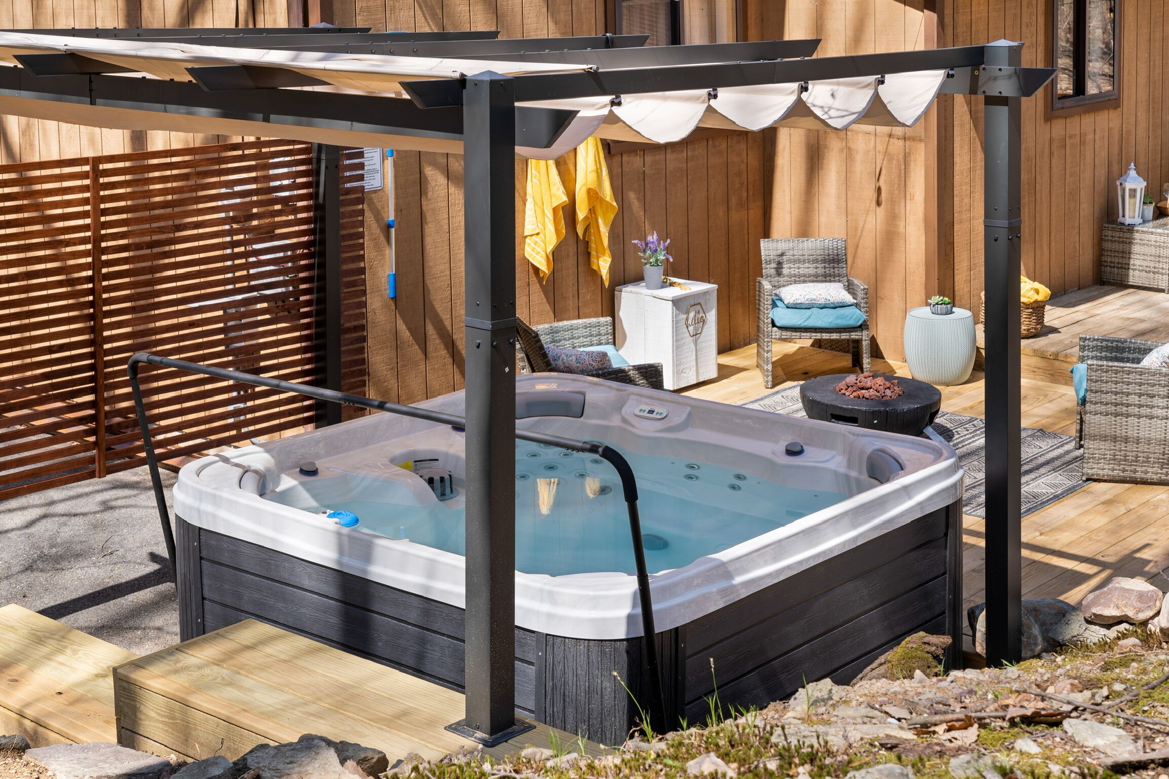 A hot tub under a pergola with a wooden deck, wicker furniture, and a stone wall in the background.