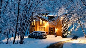 A snow-covered house with a car parked in front, surrounded by trees.