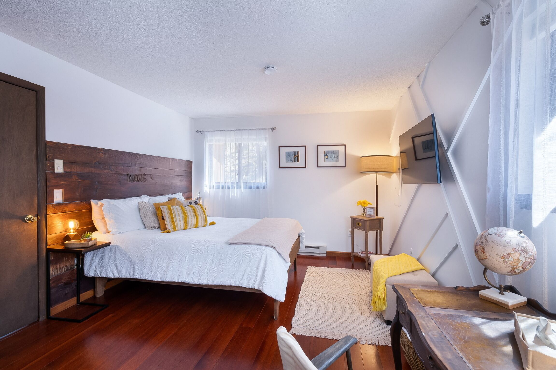 A bedroom with a wooden headboard, a bed with white bedding, a bedside table with a lamp, a globe, and a small plant.