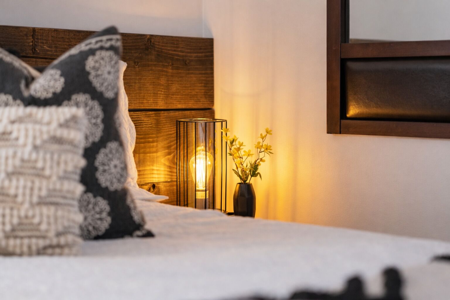 Una cama con un cojín estampado, una mesita de noche con un jarrón de flores y una lámpara fijada a la pared.