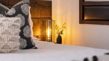 Una cama con un cojín estampado, una mesita de noche con un jarrón de flores y una lámpara fijada a la pared.