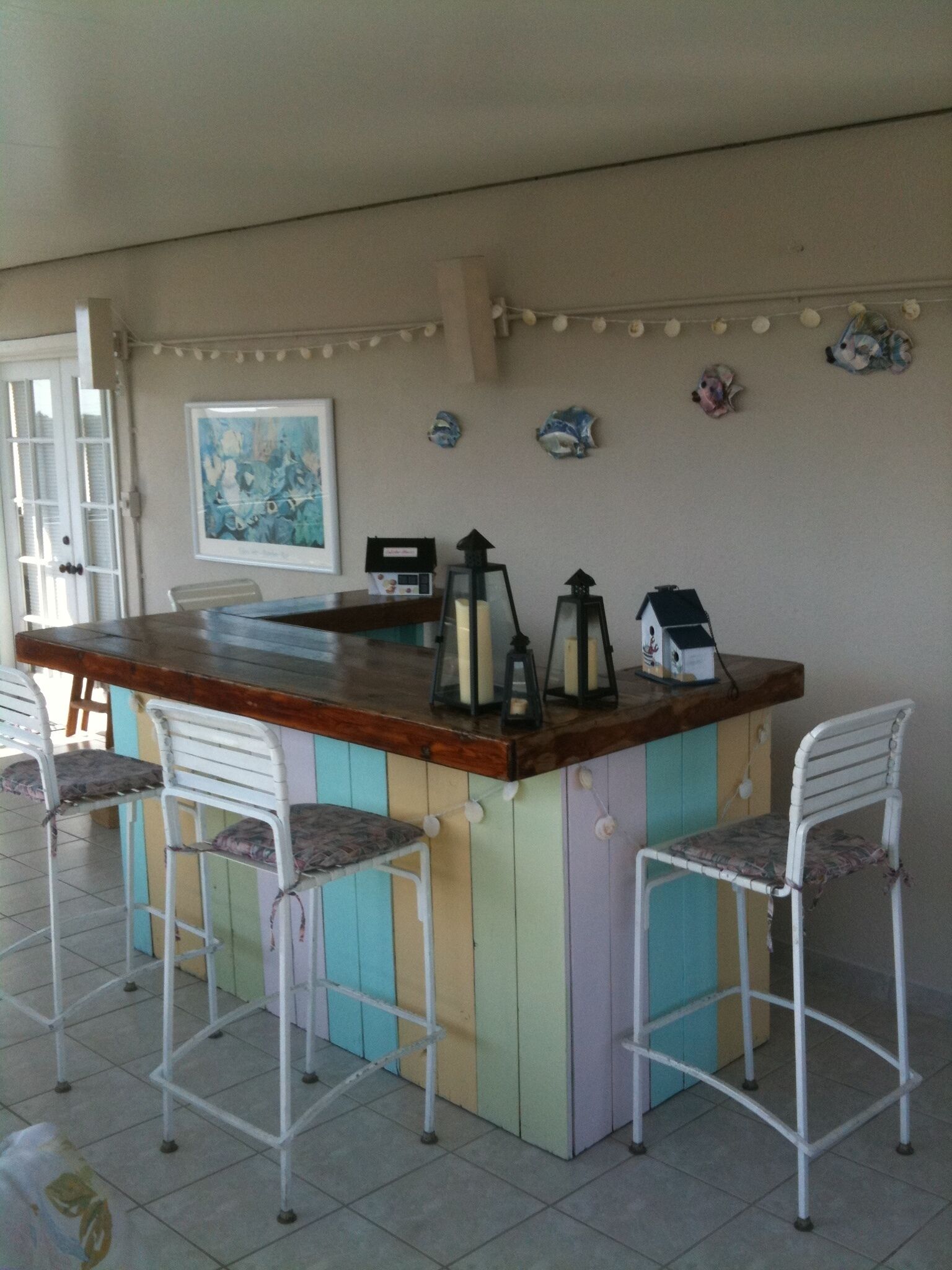 Zona bar con bancone in legno, tre sedie in metallo bianco con cuscini decorati, un'opera d'arte incorniciata a parete e oggetti decorativi come lanterne e statuine.