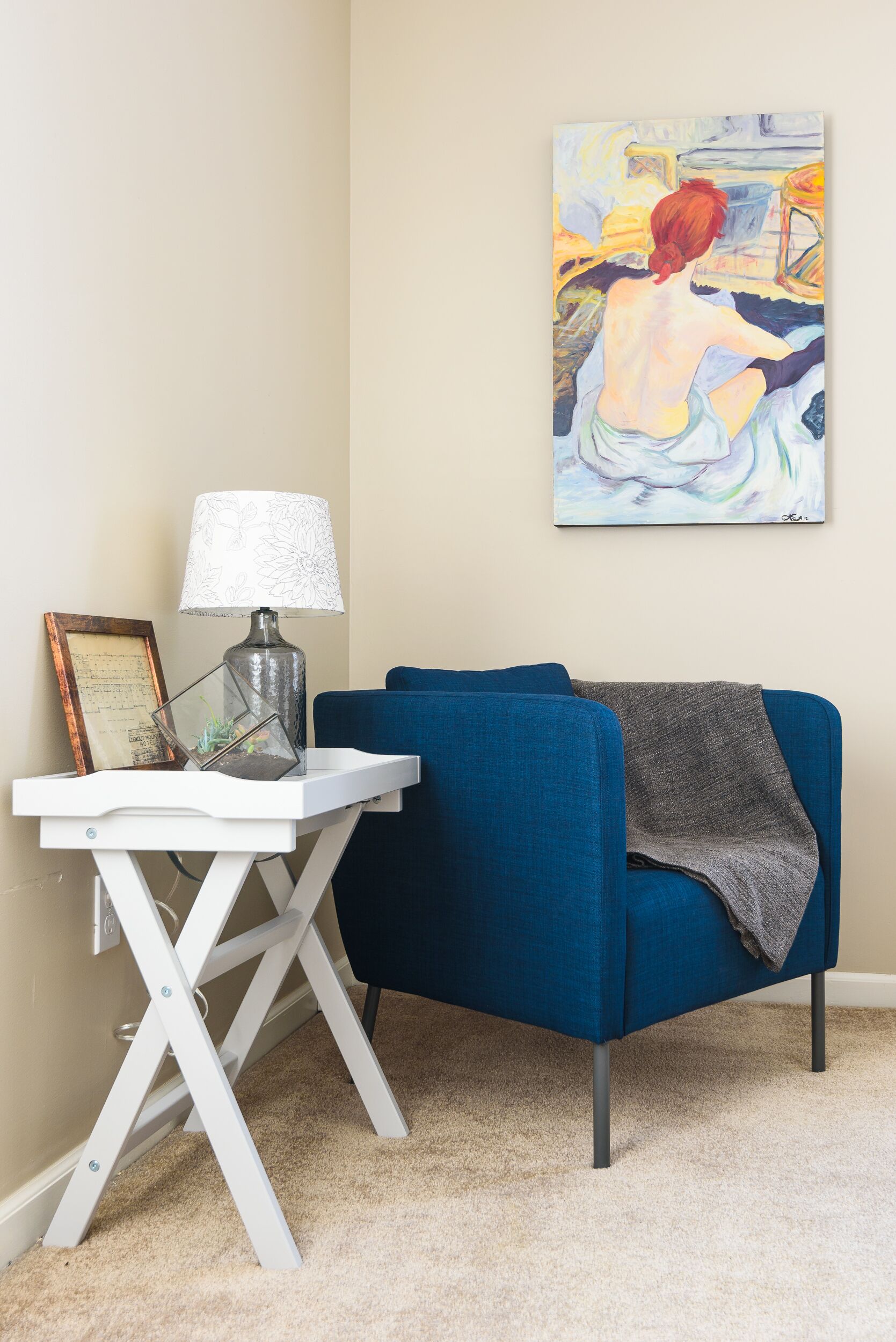Une pièce avec un fauteuil bleu, une table d’appoint blanche avec une lampe, un document encadré et un tableau accroché au mur.