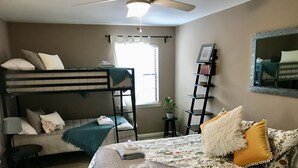 Un dormitorio con una litera, un escritorio, una silla, una mesita de noche, una lámpara, una planta en maceta, un espejo y una cama con ropa de cama y almohadas estampadas.