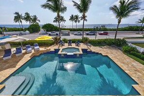 A swimming pool with lounge chairs and umbrellas, surrounded by palm trees and a view of the ocean.