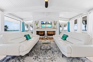 A spacious living room with white sofas, a large TV, and a fireplace.