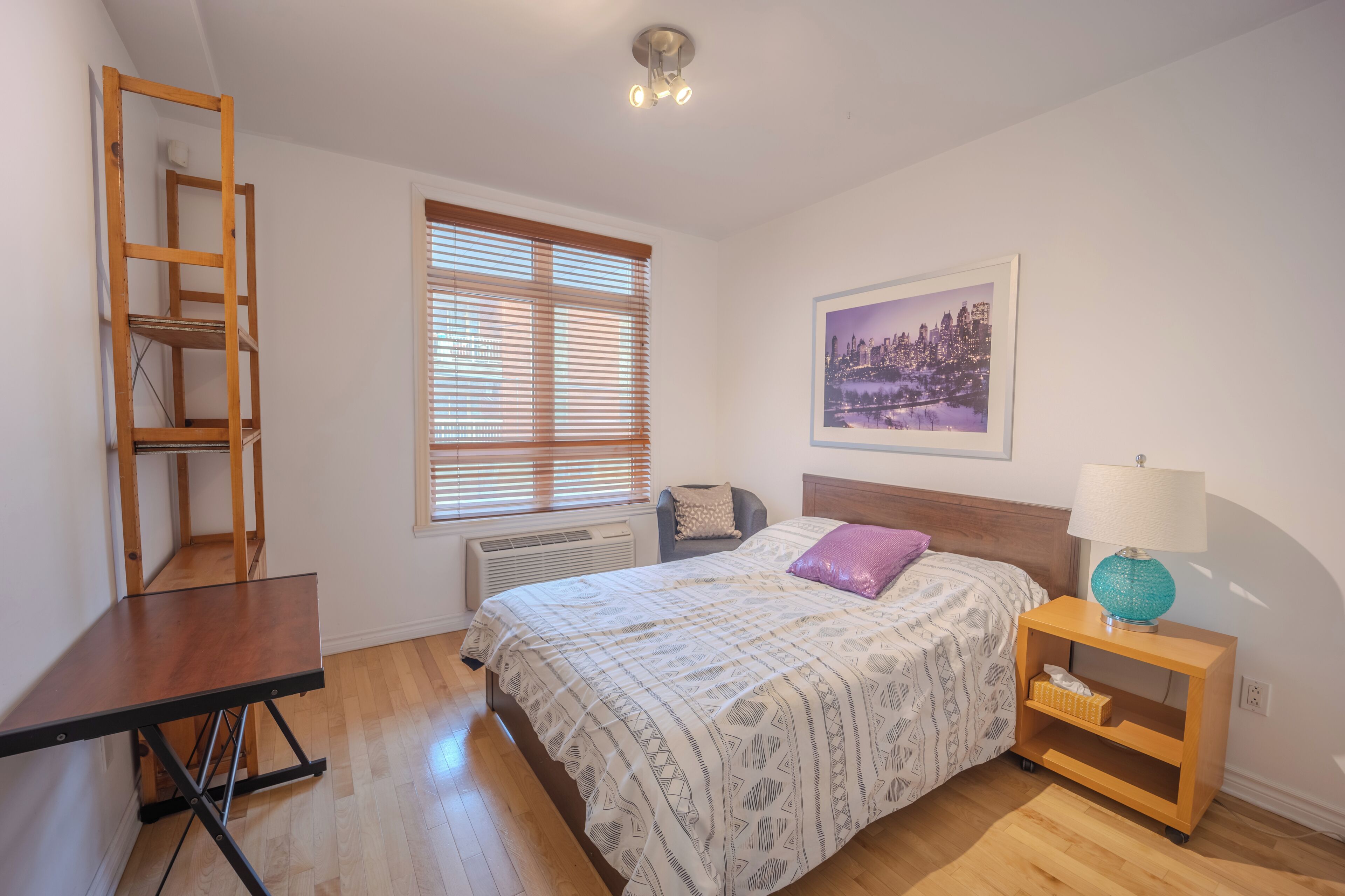 Une chambre à coucher comprenant un lit, un bureau, une chaise, une table de chevet, une lampe, une fenêtre avec des stores, un radiateur et un tableau encadré au mur.