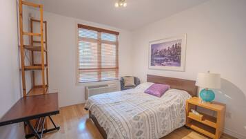 Une chambre à coucher comprenant un lit, un bureau, une chaise, une table de chevet, une lampe, une fenêtre avec des stores, un radiateur et un tableau encadré au mur.