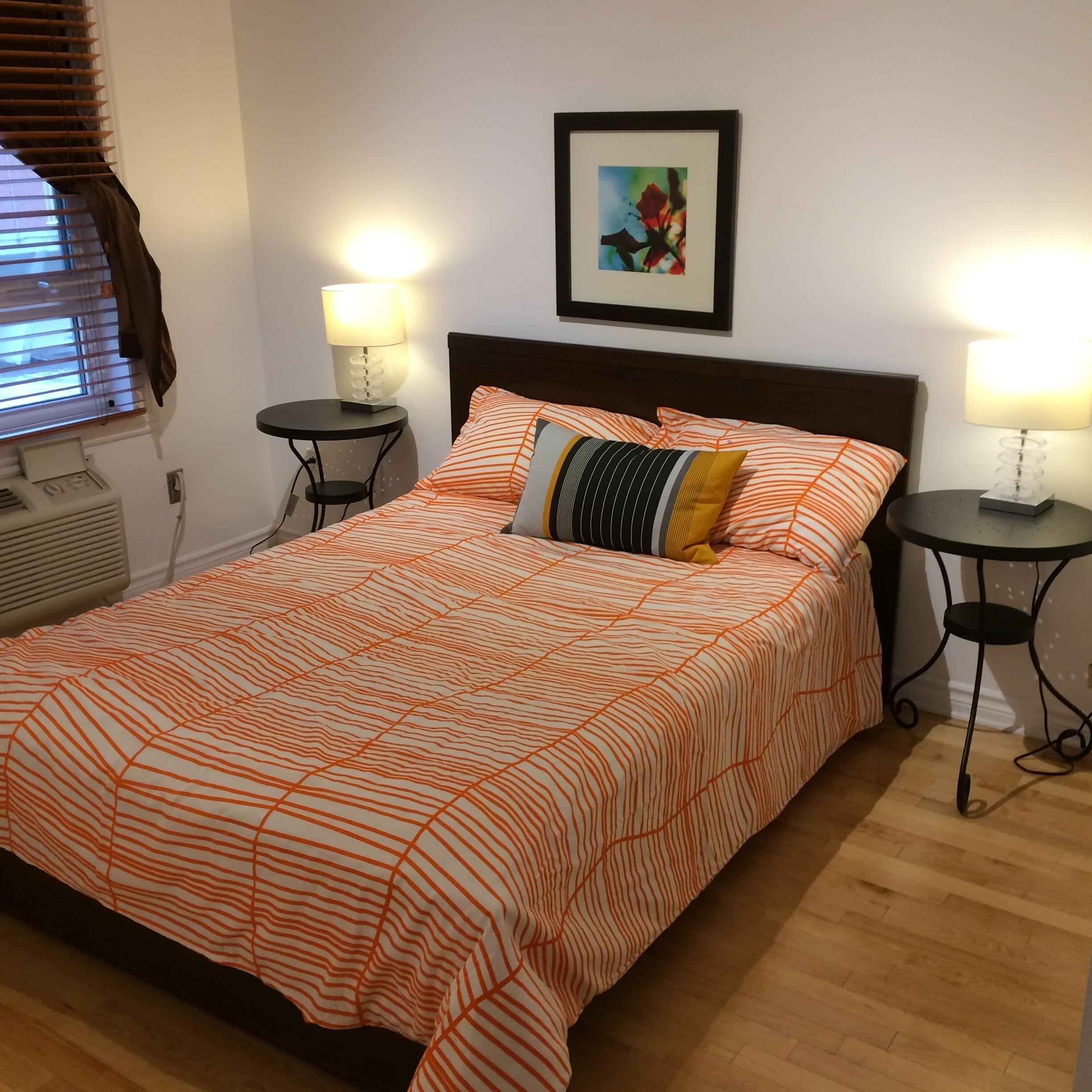 A neatly made bed with striped bedding, two bedside tables with lamps, a framed picture on the wall, and a window with blinds.