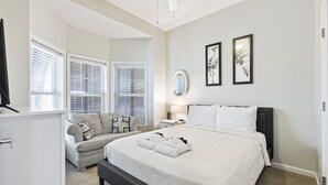 A bedroom with a large bed, a bedside table with a lamp, a dresser, and a window with blinds. - 113 Waters Edge - Sunset Haven - Renovated Riverside Townhome - Pool & Fishing Pier (Folly Beach)
