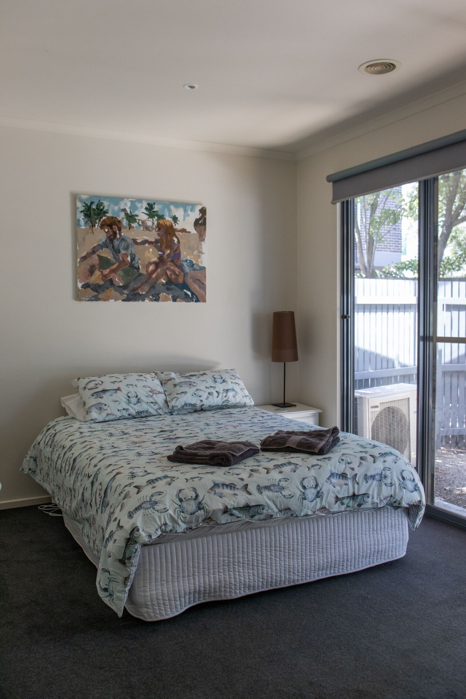 A bedroom with a large bed, a painting on the wall, a lamp, and a view through the window.