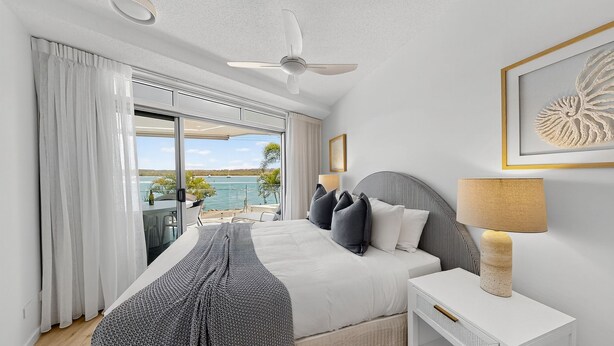 Una habitación de hotel con cama, mesita de noche, lámpara, ventilador de techo y vista al mar.