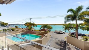 Un balcón con cerramiento de vidrio que da a una piscina, un estacionamiento y vistas al mar con barcos.