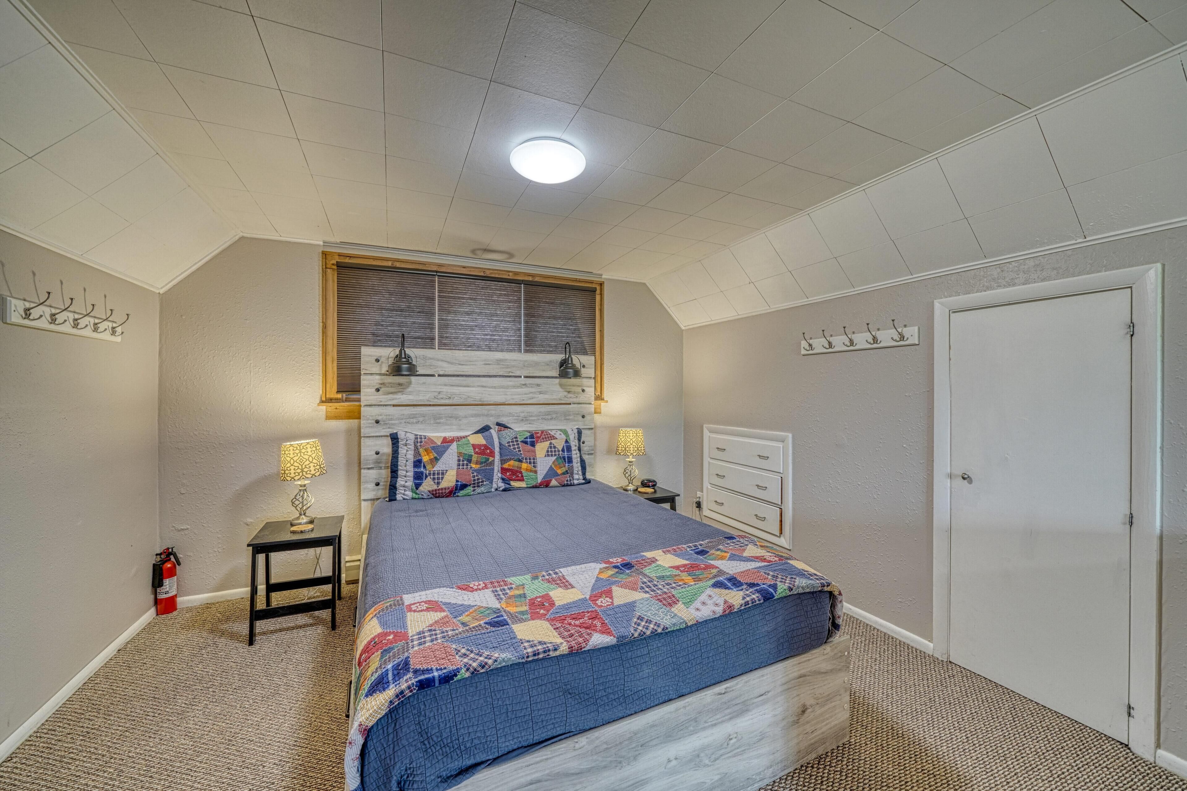 A bedroom with a bed, two bedside tables, a window, a door, and a fire extinguisher.