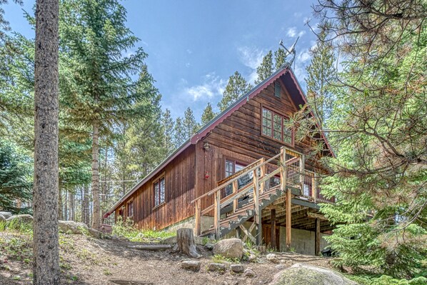 A rustic cabin with a wooden exterior, a staircase leading to a deck, surrounded by dense forest.
