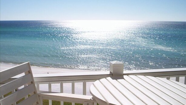 Ein Balkon mit weißer Brüstung und einem Tisch, der Blick auf einen Strand mit klarem, blauem Wasser und der hellen Sonne bietet, die sich auf der Oberfläche spiegelt.
