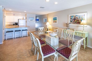 A dining area with a glass table and white chairs, a kitchen in the background, and a painting on the wall.