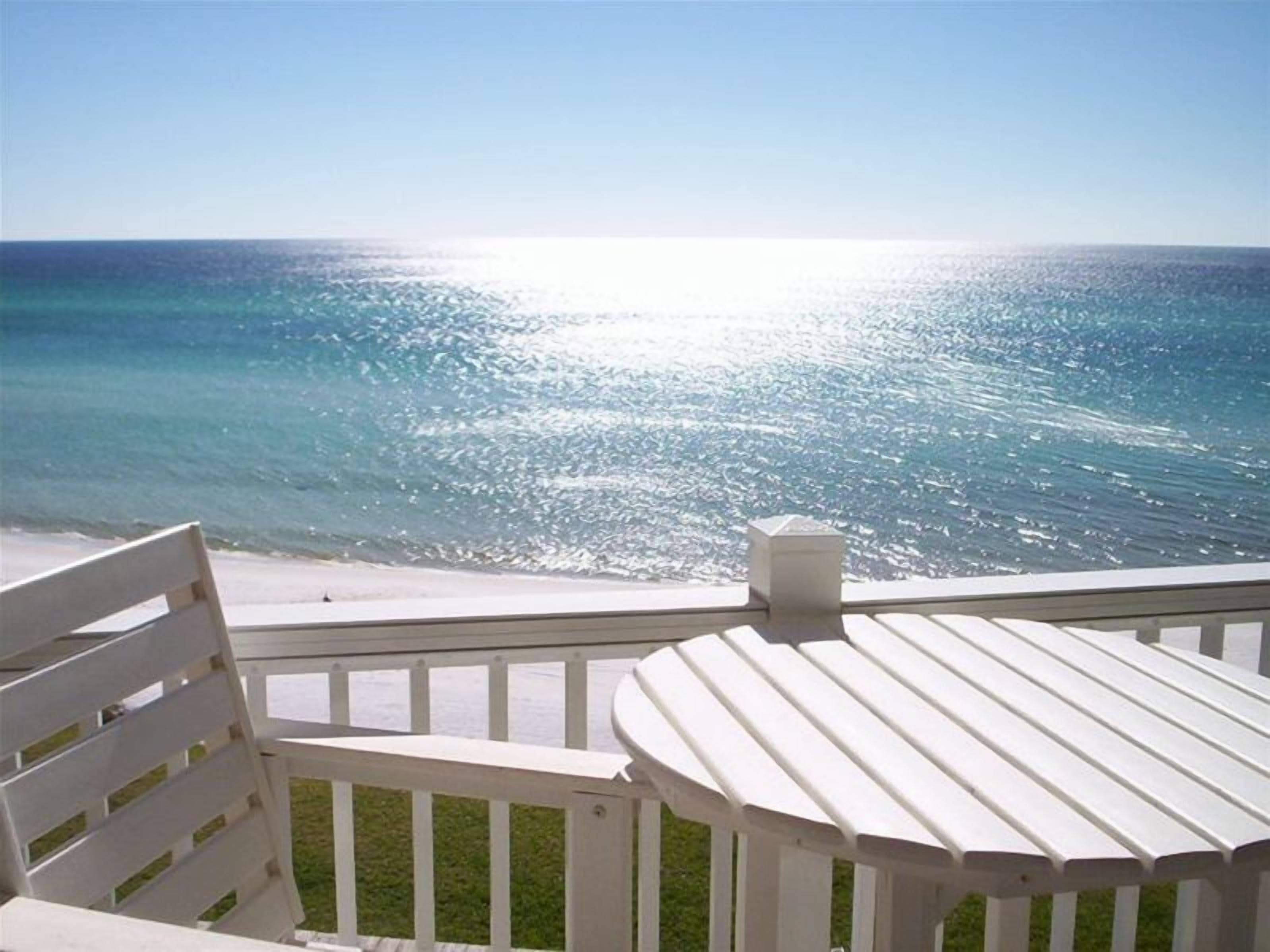 Un balcon doté d’une balustrade blanche et d’une table, offrant une vue sur une plage aux eaux d’un bleu limpide et sur un soleil éclatant qui se reflète à la surface de l’eau.