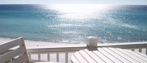 Un balcone con ringhiera bianca e un tavolo, con vista su una spiaggia e acqua cristallina, con il sole che si riflette sulla superficie.
