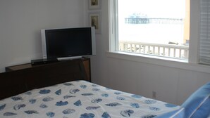 A bedroom with a bed, a TV on a stand, a window with a view of a balcony, and a wall with framed pictures. - Exclusive Oceanfront Cottage on Dry Sandy Carbon Beach - Seal (Malibu)