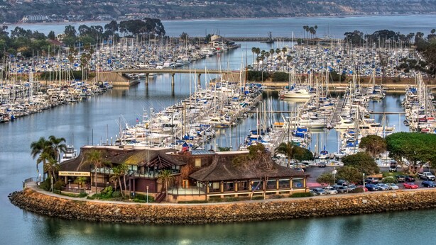 Uma marina com vários barcos atracados, um restaurante chamado "DANA PRINT VINTAGE CLUB" à esquerda e uma ponte ao fundo.