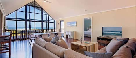 A spacious living area with a large window overlooking the sea, a comfortable sofa, a dining table, and a flat-screen TV.