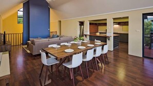 A dining area with a wooden table set for six, a sofa, and a kitchen in the background.