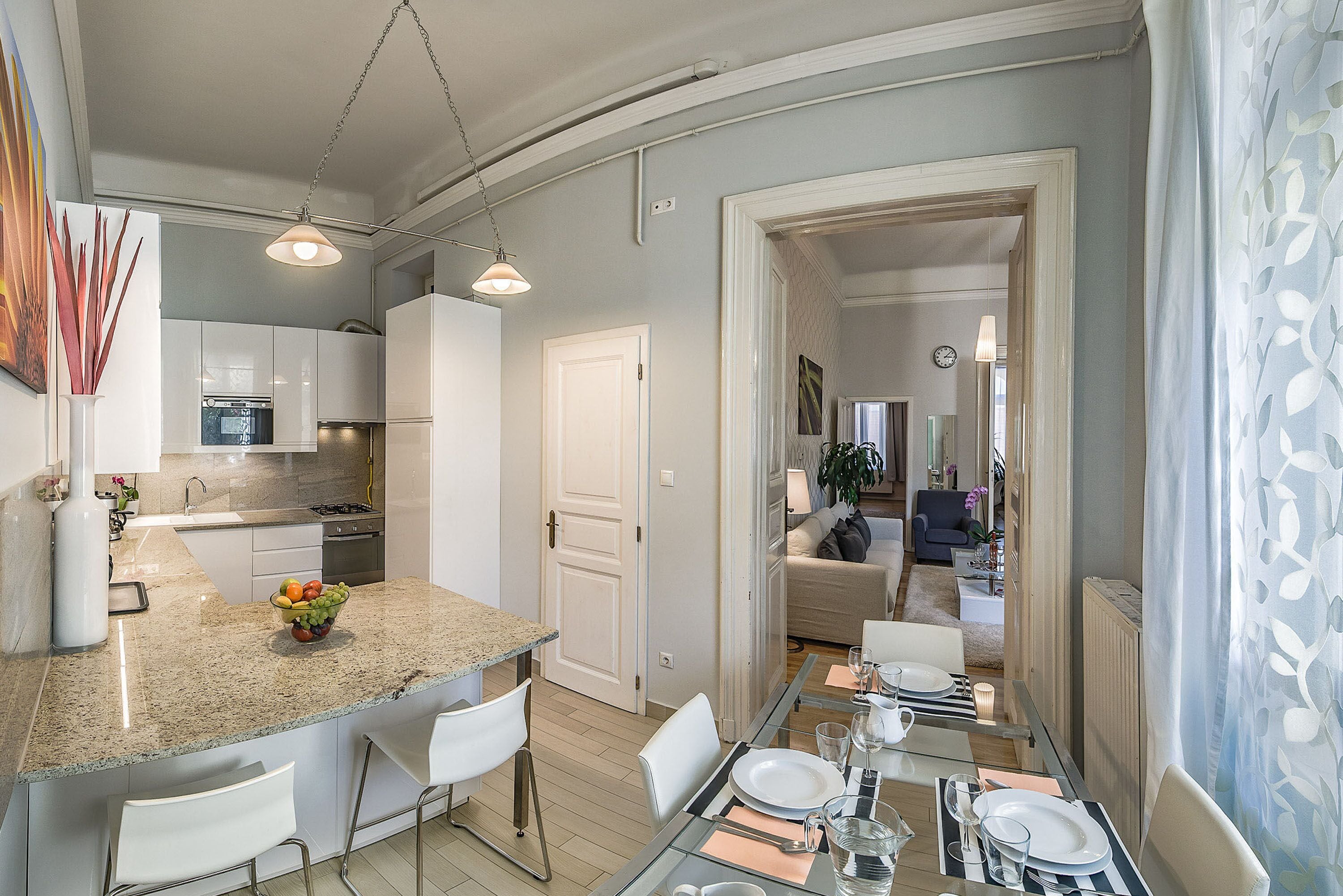 A modern kitchen with a central island, dining area with a glass table, and a living space visible through an open door.