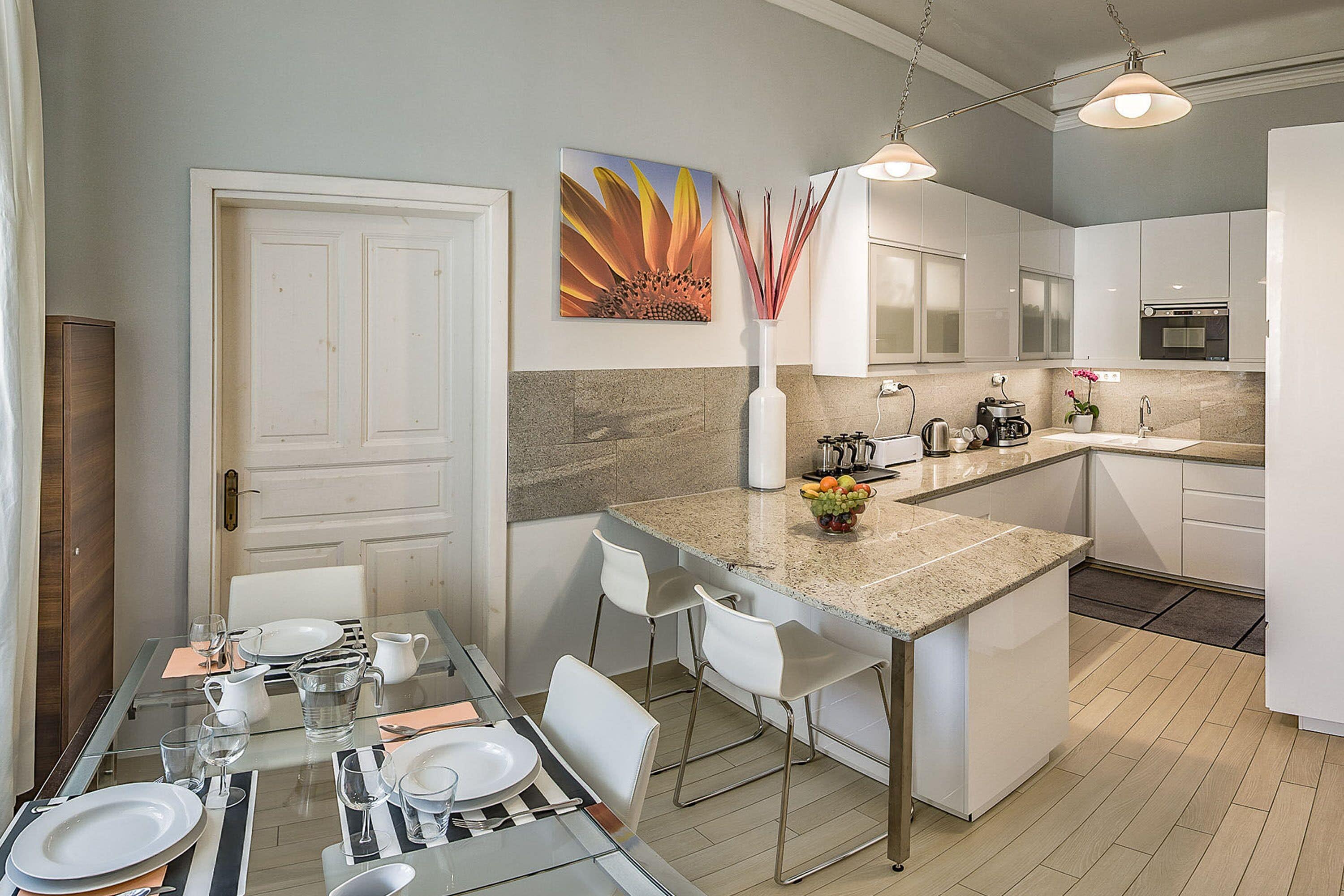 Una cocina moderna con armarios blancos, encimera de granito y comedor con mesa de vidrio y sillas blancas.