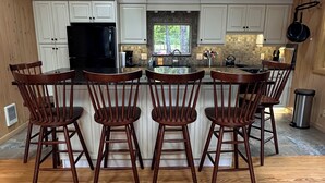 A kitchen with wooden cabinets, a central island with bar stools, hanging pendant lights, and a window overlooking greenery. - Swim and Kayak at this freshwater location. Sleep 6+ on MDI near Acadia National (Seal Cove)
