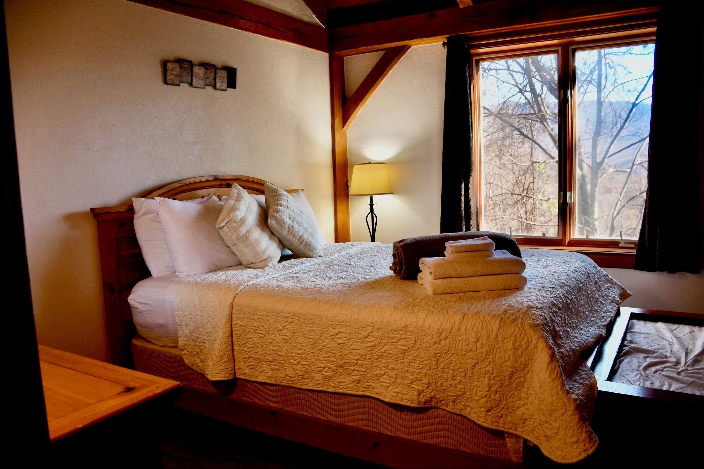 Un dormitorio con cama, almohadas, una mesita de noche, lámpara y ventana con vistas.