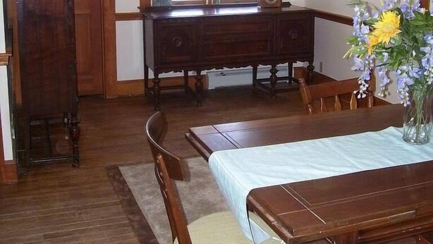 Un comedor con una mesa y sillas de madera, una ventana con cortinas y un jarrón con flores sobre la mesa.