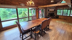 A wooden interior with a dining table, chairs, a chandelier, and large windows offering a view of the outdoors.