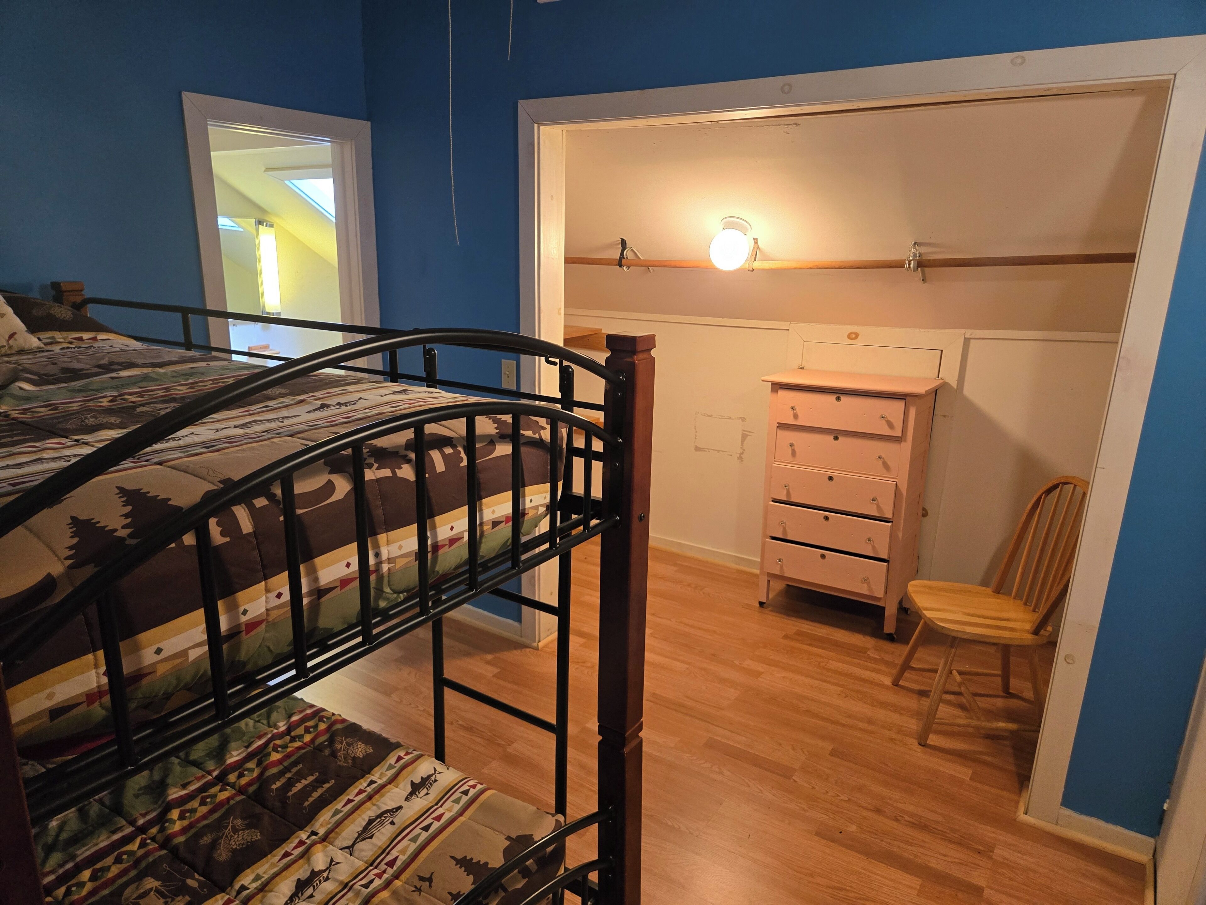 Ein Zimmer mit einem Etagenbett, einer Holzkommode, einem Stuhl und einer Tür zu einem anderen Raum.