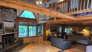 A spacious living area with a fireplace, wooden flooring, a sofa, a dining table, and large windows offering a view of the outdoors.