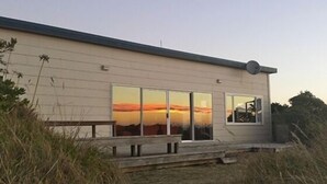 A single-story building with large windows reflecting a sunset. - Magical Beach Kiwi Bach (Waitarere Beach)