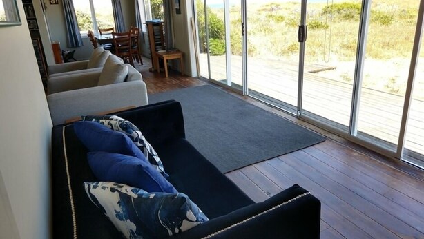 A spacious living area with a sofa, a dining table, and a view of the beach through large sliding glass doors. - Magical Beach Kiwi Bach (Waitarere Beach)