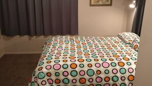 A bedroom with a bed covered in a polka dot bedspread, a window with curtains, and a framed picture on the wall. - Magical Beach Kiwi Bach (Waitarere Beach)